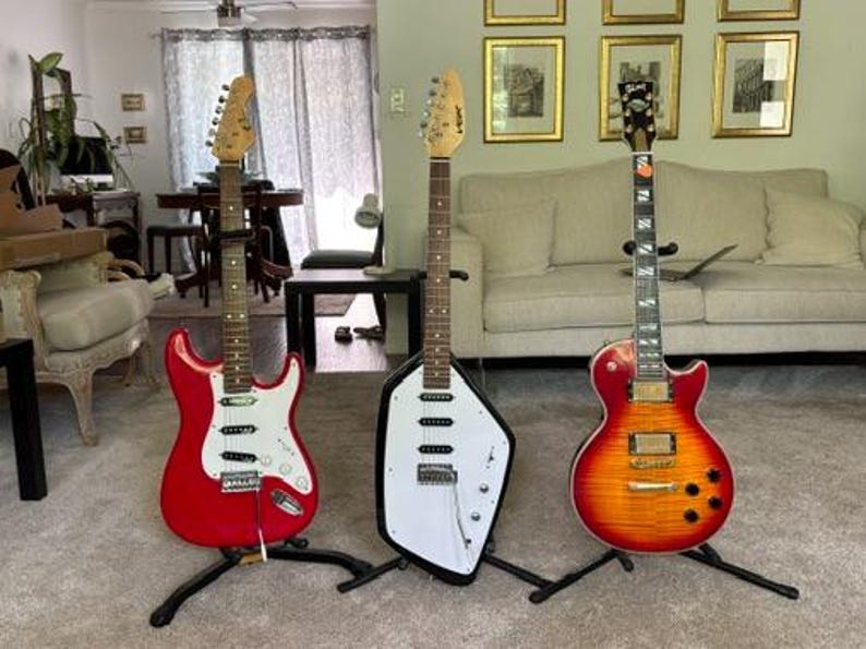 May include: Three electric guitars standing on individual black stands. The left is a red double-cutaway guitar with a white pickguard. The center is a white, angular, asymmetrical guitar. The right is a sunburst finish single-cutaway guitar.