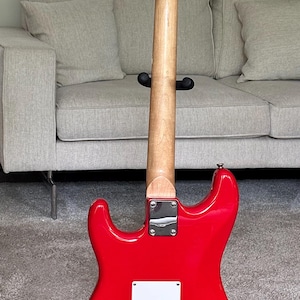 May include: A bright red electric guitar shown from the back, featuring a smooth glossy finish, a light-colored wooden neck, a chrome neck plate, and a white rectangular back plate with six string holes. The headstock includes silver tuning pegs.