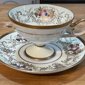 Può includere: Un set da tè bianco con bordo dorato e un intricato motivo floreale in oro e rosa. La tazza ha una base a piedistallo e un manico delicato.