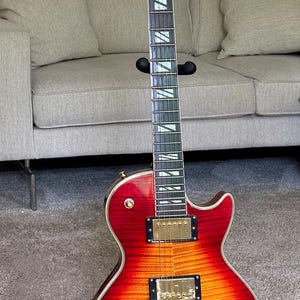 May include: A Gibson electric guitar with a cherry sunburst finish on a flamed maple top. The instrument features gold hardware, two humbucker pickups, a tune-o-matic bridge, four black control knobs, and a dark fretboard with trapezoid inlays.
