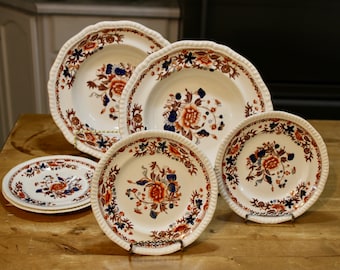 Op de afbeelding: Een verzameling antieke witte keramische borden met een bloemmotief in oranje, blauw en bruin. De borden hebben een geschulpte rand en worden op een houten oppervlak gepresenteerd.
