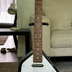May include: A Vox electric guitar with a distinctive teardrop-shaped body, featuring a white pickguard, a black outer frame, and a wooden neck with a dark fretboard. The headstock displays the Vox logo, and the body includes three pickups.