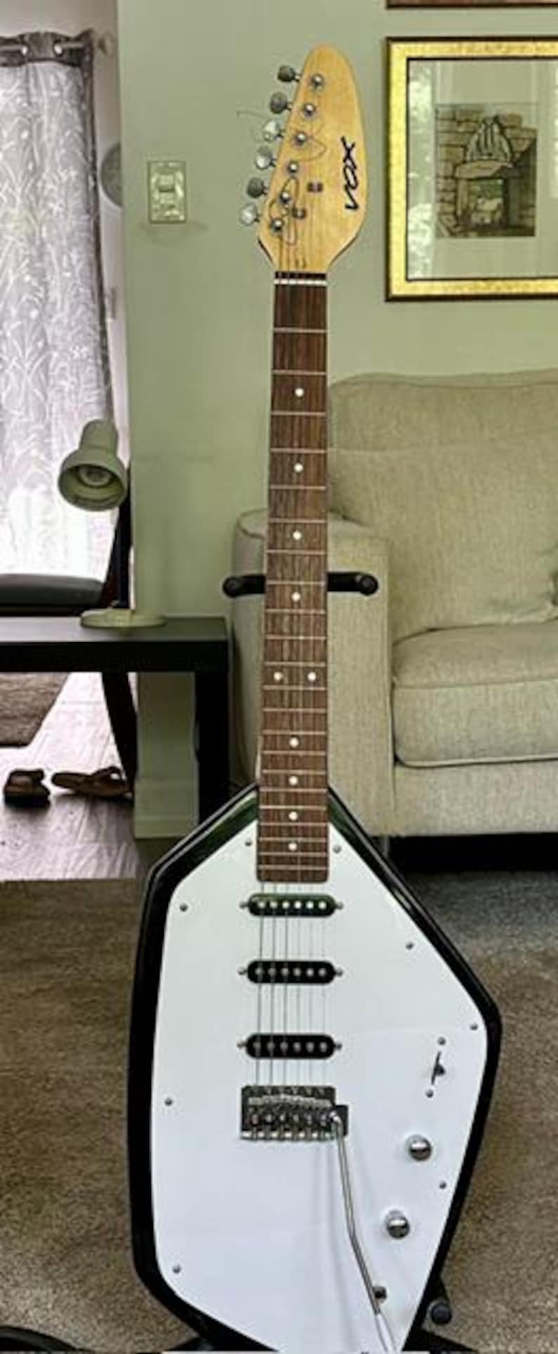 May include: A Vox electric guitar with a distinctive teardrop-shaped body, featuring a white pickguard, a black outer frame, and a wooden neck with a dark fretboard. The headstock displays the Vox logo, and the body includes three pickups.