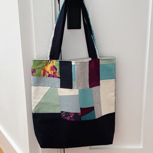 May include: A patchwork tote bag with a black base and colorful fabric squares. The bag has two long straps and is hanging on a white door.