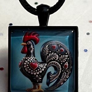 May include: Square pendant necklace featuring a stylized black rooster with red accents and heart details. The rooster has a red comb and yellow feet. The pendant hangs from a black cord.