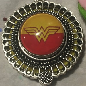 Puede incluir: Un colgante de plata con un logotipo de Wonder Woman amarillo y rojo en un cabujón redondo de vidrio. El colgante está rodeado por un marco decorativo de plata.