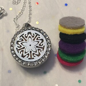 May include: Silver-toned aromatherapy necklace with a decorative pendant featuring a floral design and clear crystal accents. A stack of colorful, round felt pads in shades of gray, yellow, black, purple, green, and pink are next to the necklace.