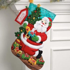 May include: A felt Christmas stocking featuring a Santa Claus figure in a red suit with white trim, a green Christmas tree, and a toy train. The stocking has a red tag with the name Stan embroidered in white, decorated with sequins and beads.