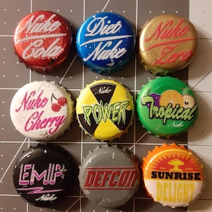 May include: Nine colorful bottle caps with various designs and text. The caps feature the words "Nuke Cola", "Diet Nuke", "Nuke Zero", "Nuke Cherry", "Nuke Power", "Tropical", "EMIP", "DEFCON", and "Sunrise Delight".