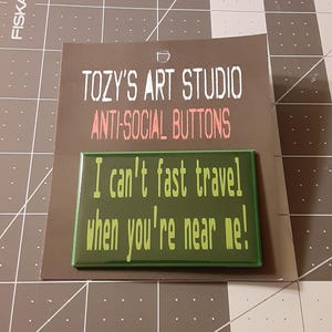 May include: A rectangular green button with the text "I can't fast travel when you're near me!" is displayed on a brown card. The card also features the text "TOZY'S ART STUDIO" and "ANTI-SOCIAL BUTTONS" in white and pink.