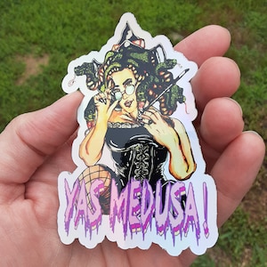 May include: A holographic sticker featuring a cartoon illustration of Medusa with snakes for hair, wearing a black corset and fishnet stockings. The sticker has a purple dripping text that says "Yas Medusa!"