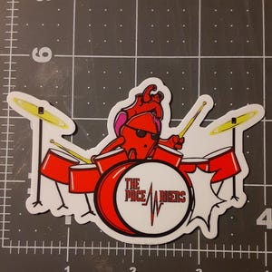 May include: A sticker depicting a red heart character wearing sunglasses, playing a red and white drum kit with yellow cymbals. The bass drum has the text "THE PACE RIKERS" in a circular design.