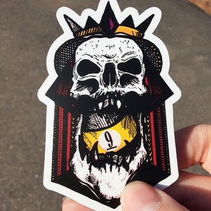 May include: A black and white sticker of a skull wearing a crown and holding a yellow pool ball with the number 9 on it.
