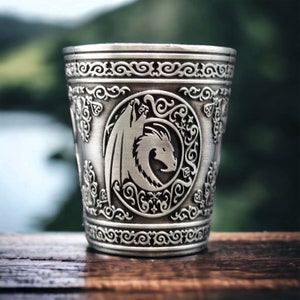 May include: A silver metal cup with an embossed design of a dragon in a circle. The cup has a decorative border around the top.