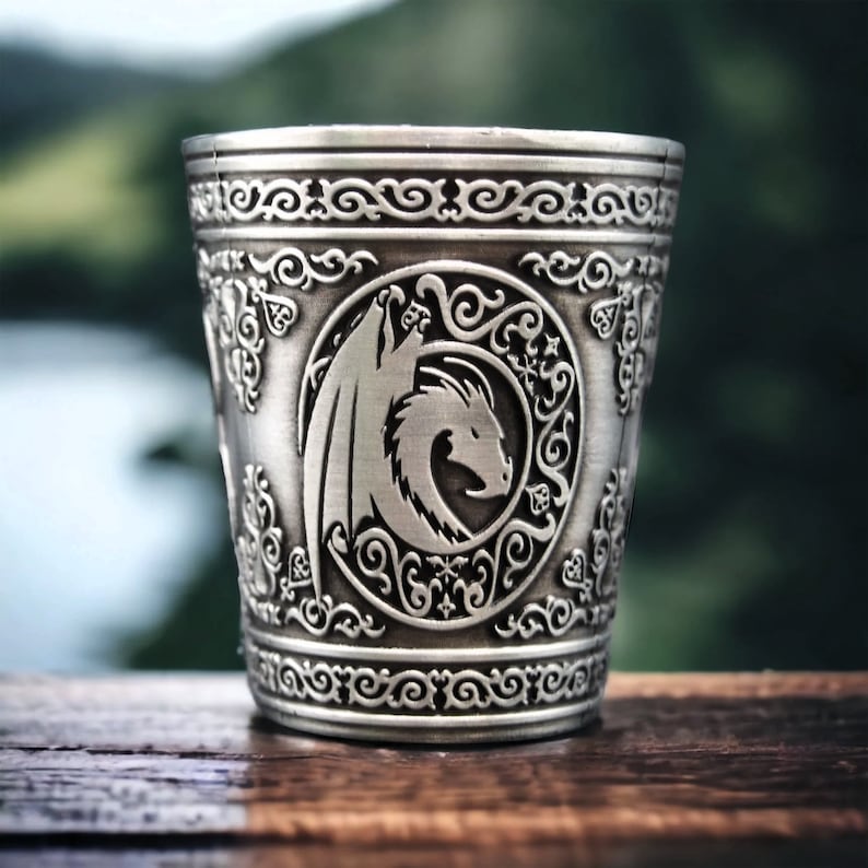 May include: A silver metal cup with an embossed design of a dragon in a circle. The cup has a decorative border around the top.