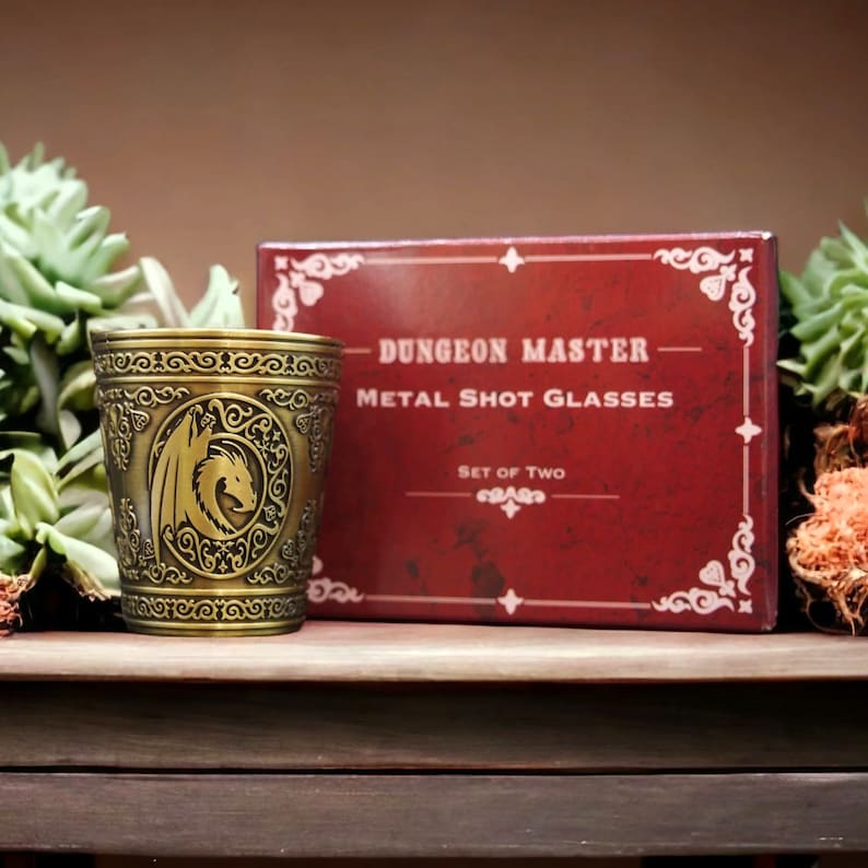 May include: A set of two gold metal shot glasses with a dragon design. The glasses are in a red box with the text "Dungeon Master Metal Shot Glasses - Set of Two".