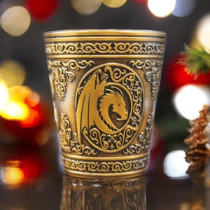 May include: A gold metal drinking cup with an ornate design featuring a dragon. The cup has a raised rim and a detailed pattern around the top and bottom.