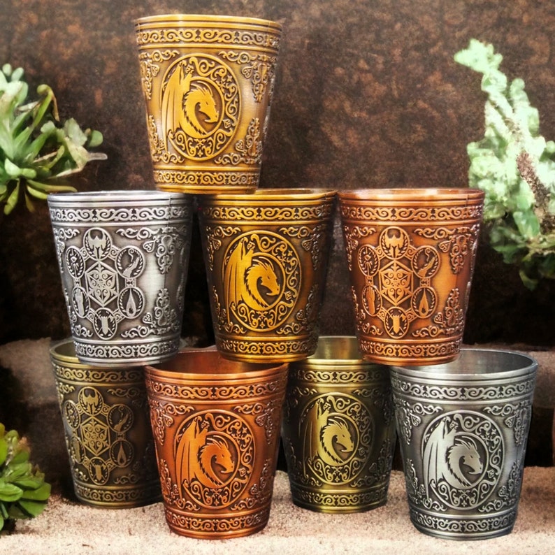 May include: Eight metal shot glasses with different designs. Each glass features a dragon, a geometric pattern, or a combination of both. The glasses are in various shades of gold, silver, and copper.