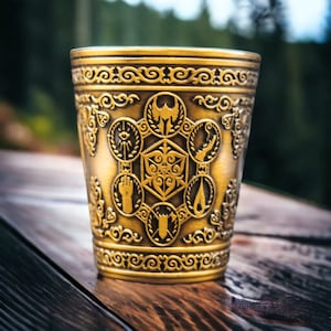 May include: A gold-colored metal cup with an intricate design featuring various symbols, including a five-pointed star, a hand, a flame, and a dice.
