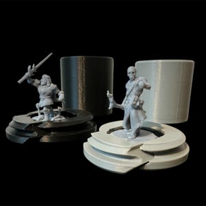 May include: Two 3D printed miniature figures, one with a sword and the other with a staff, stand on separate bases. The bases are made of a layered material, one black and one grey.