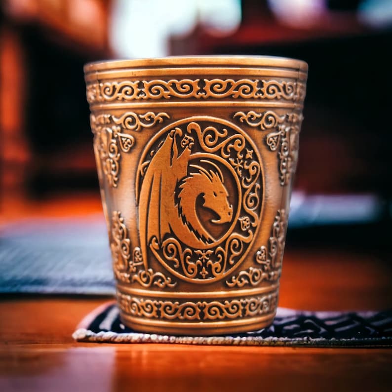May include: A gold-colored metal cup with an ornate design featuring a dragon in a circle. The cup has a textured surface and a wide rim.
