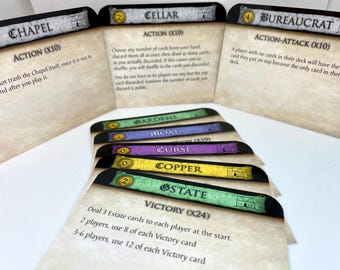 May include: A set of Dominion card game cards, including Chapel, Cellar, Bureaucrat, Gardens, Moat, Curse, Copper, and Estate. The cards are color-coded and feature illustrations of castles and other medieval imagery.