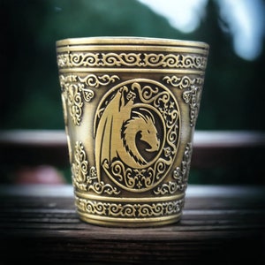 May include: A gold-colored metal shot glass with an ornate design featuring a dragon. The dragon is in a circular frame with intricate details.