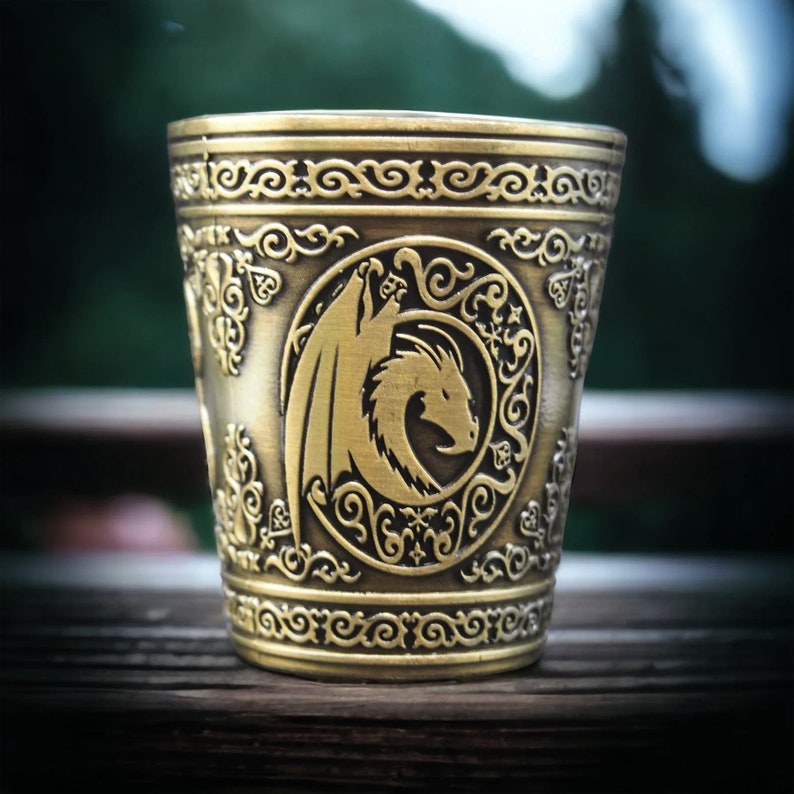 May include: A gold-colored metal shot glass with an ornate design featuring a dragon. The dragon is in a circular frame with intricate details.