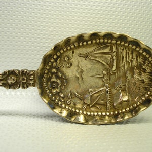 May include: A silver spoon with an ornate handle and a detailed engraved design of a windmill and a sailboat on the bowl.