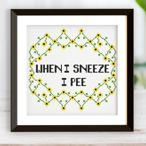 May include: Framed cross-stitch artwork. The design features the text "WHEN I SNEEZE I PEE" in black, surrounded by a border of green leaves and yellow flowers. The frame is black and rectangular, displayed on a wooden surface.