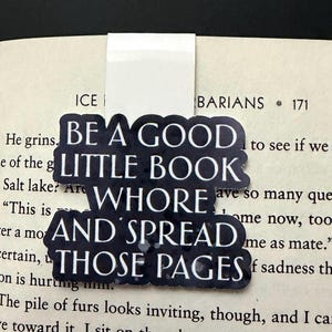 May include: A magnetic bookmark featuring a dark, textured background with white text that reads: BE A GOOD LITTLE BOOK WHORE AND SPREAD THOSE PAGES. The bookmark is clipped onto the top edge of an open book page with visible printed text.
