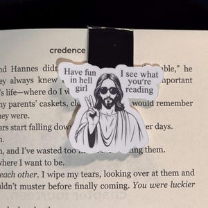May include: A white bookmark with a black and white illustration of Jesus giving a peace sign. The text on the bookmark reads "Have fun in hell girl" and "I see what you're reading".