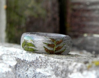 May include: A clear resin ring with embedded green and brown leaves. The ring is sitting on a gray, textured surface.