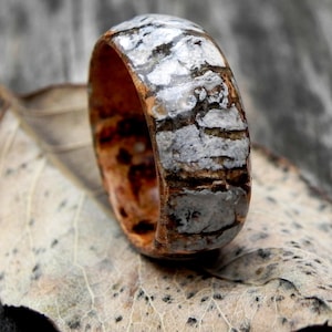 May include: A wooden ring with a distinctive pattern of brown, grey, and white. The ring is displayed on a dried leaf, emphasizing its natural appearance. The close-up shot reveals the ring's detailed craftsmanship. The ring's design resembles natural wood grain.