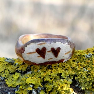 May include: A wooden ring with a clear resin coating, featuring two brown hearts carved into the wood. The ring is resting on a bed of green moss.