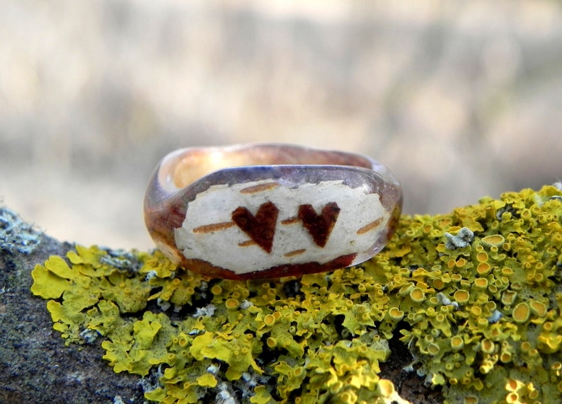 May include: A wooden ring with a clear resin coating, featuring two brown hearts carved into the wood. The ring is resting on a bed of green moss.