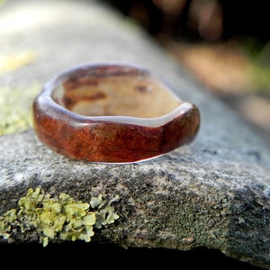 May include: A close-up of a wooden ring with a reddish-brown hue and a clear resin finish. The ring is resting on a textured, grey surface.