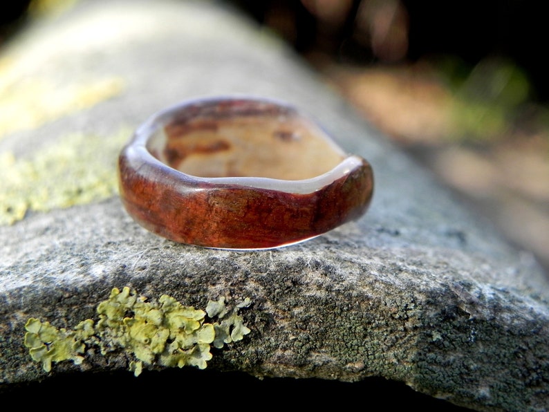 May include: A close-up of a wooden ring with a reddish-brown hue and a clear resin finish. The ring is resting on a textured, grey surface.