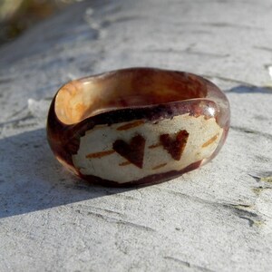 May include: A brown resin ring with two heart-shaped designs on the top. The hearts are made of a lighter brown material and are set against a white background.