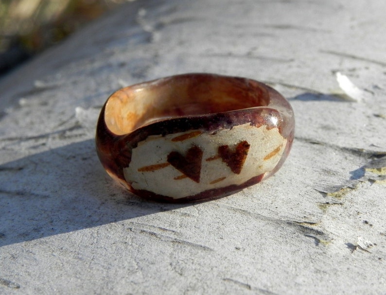 May include: A brown resin ring with two heart-shaped designs on the top. The hearts are made of a lighter brown material and are set against a white background.