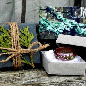 May include: A wooden ring with a clear resin inlay featuring pink flowers. The ring is in a small grey box with white tissue paper. The box is on a wooden surface. The text "Forest for you" is on a card in the background.