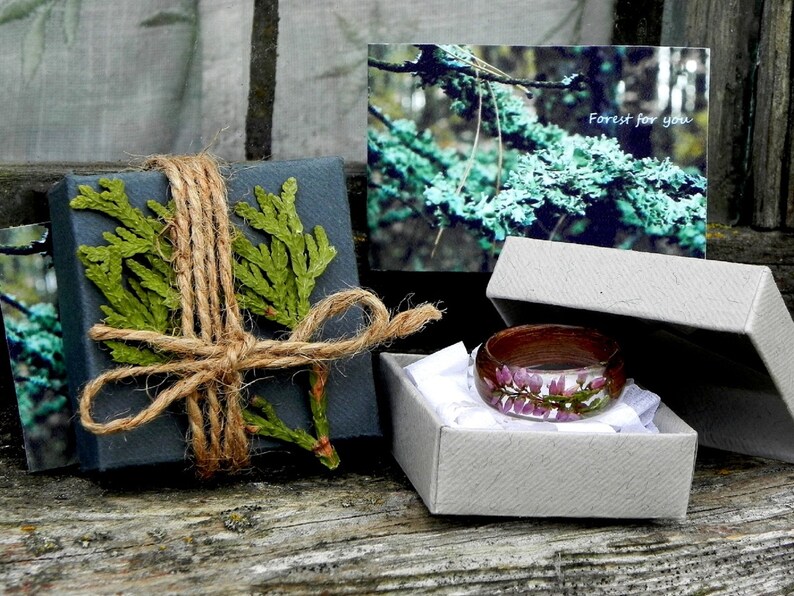 May include: A wooden ring with a clear resin inlay featuring pink flowers. The ring is in a small grey box with white tissue paper. The box is on a wooden surface. The text "Forest for you" is on a card in the background.