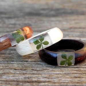 May include: Three rings made of wood and clear resin, each featuring a single embedded four-leaf clover. One ring is light brown wood, one is white wood, and one is dark brown wood. The bands are smooth and circular in shape.