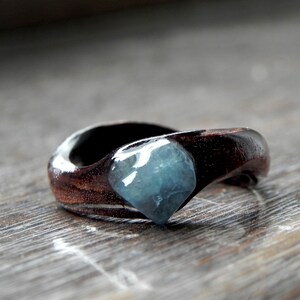 May include: A handcrafted wooden ring with a dark brown finish. The ring features a light blue, gem-like stone set into the wood. The ring is a unique piece of jewellery.