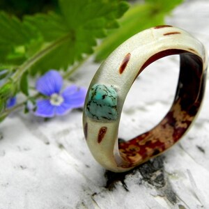 Könnte beinhalten: Ein weißer und brauner Holzring mit einem türkisfarbenen Stein-Inlay. Der Ring liegt auf einem weißen Hintergrund mit grünen Blättern.