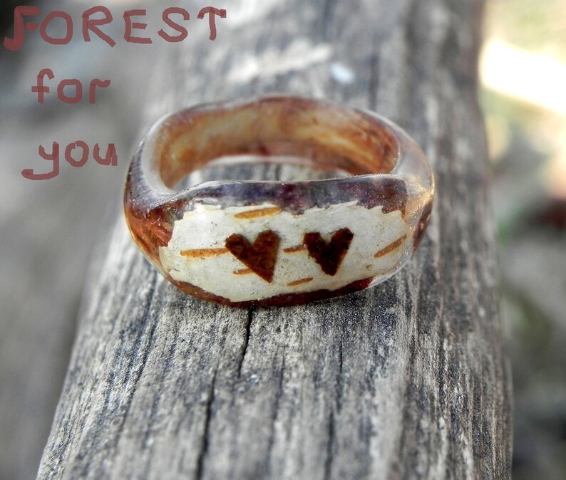 May include: A clear resin ring with a wood-like texture and two brown hearts. The text "FOREST for you" is visible in the background.