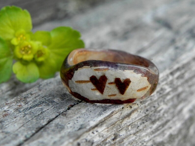 May include: A wooden ring with a clear resin coating. The ring has two hearts carved into the wood, and the hearts are painted brown. The ring is sitting on a piece of weathered wood.