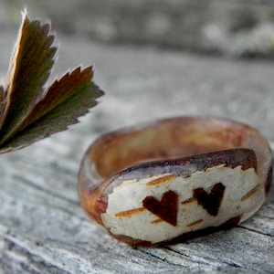 May include: A wooden ring with a white background and two brown hearts carved into it. The ring is sitting on a piece of weathered wood.