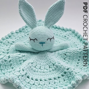 May include: A crocheted baby security blanket in a soft, light blue color, featuring a bunny head with closed eyes and long ears. The blanket has a textured, scalloped edge and the text "PDF CROCHET PATTERN" in white.