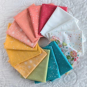 May include: A collection of folded fabric squares in a circular pattern, showcasing a range of colours such as coral, yellow, and teal. Some fabrics display floral and dot patterns. The fabrics are arranged on a white, quilted surface.